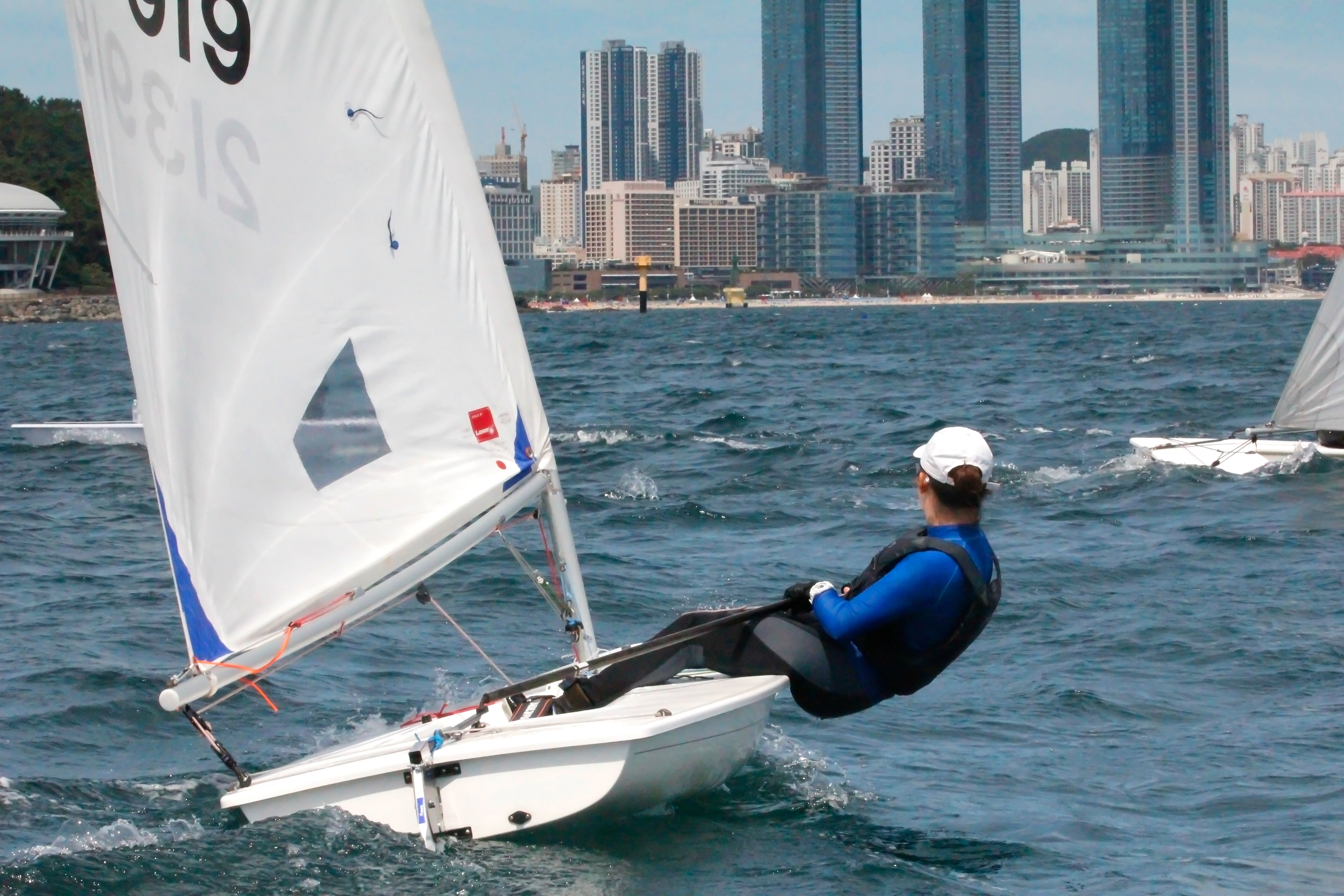 Person sailing a small boat on the water with city buildings in the background during Son Jiwon yachting journey